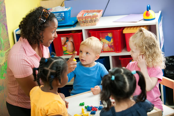 a female teacher talking to four small children with a toy shelf in the background